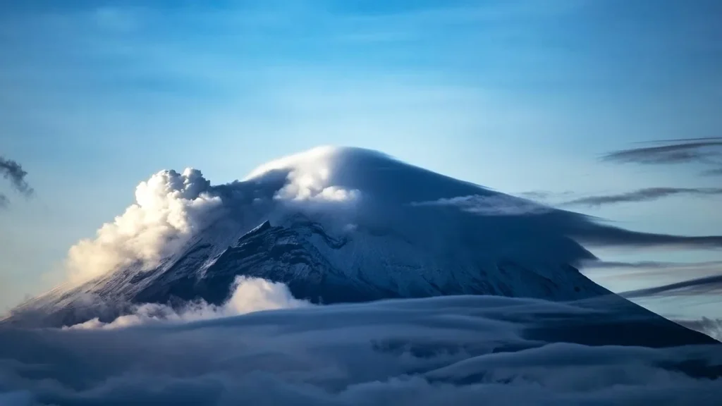 A majestic, snow-capped volcanic peak rises through a thick layer of clouds under a blue sky.