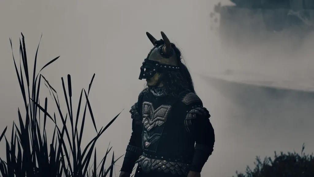 A Viking warrior in dark armor stands in a foggy marsh with tall reeds.