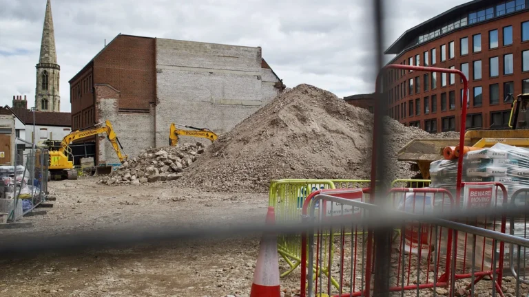 Construction site with a large pile of dirt, yellow excavators, and safety barriers behind a blurry fence.