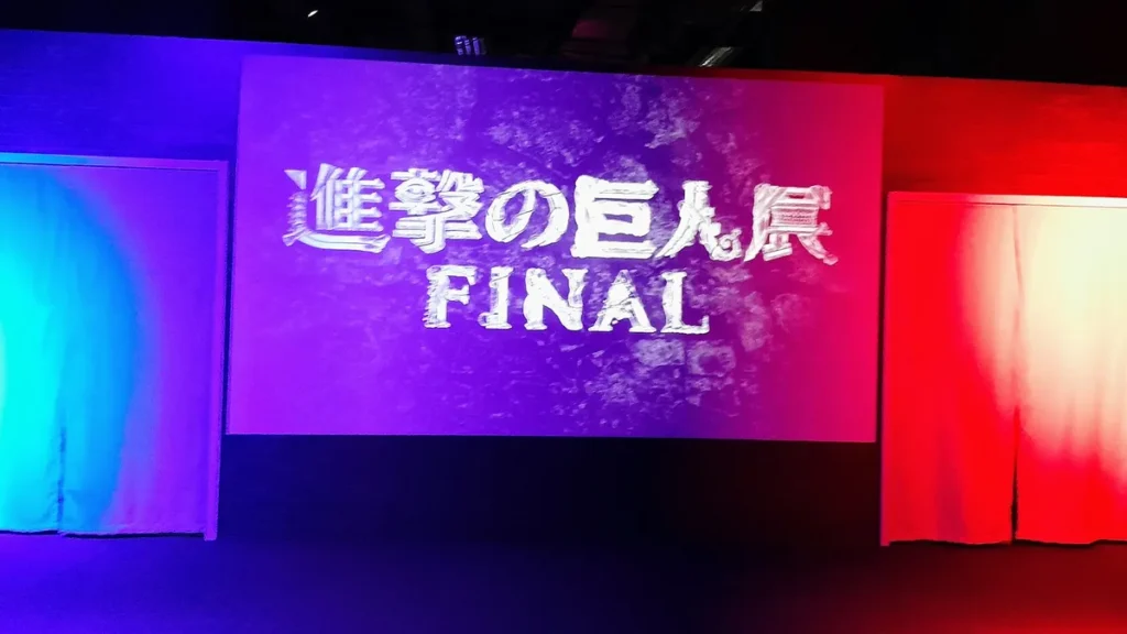 Colorful stage screen showing the stylized Attack on Titan Exhibition FINAL logo in Japanese and English.