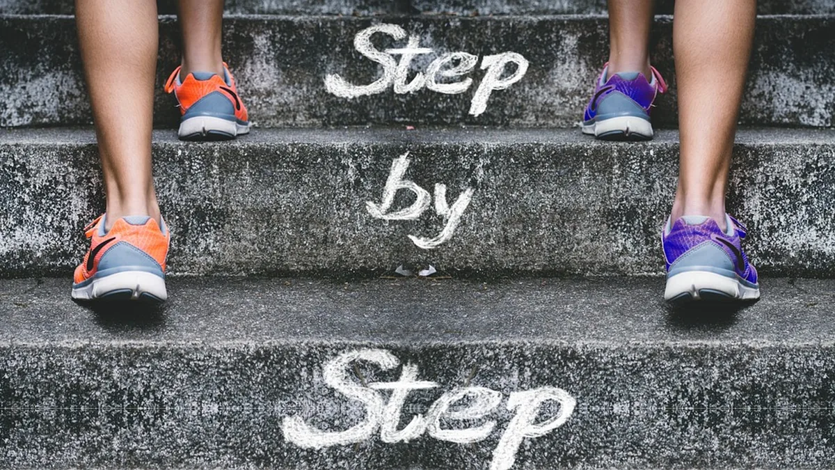 Legs in orange and purple sneakers stand on concrete steps with 'Step by Step' written in white chalk text.