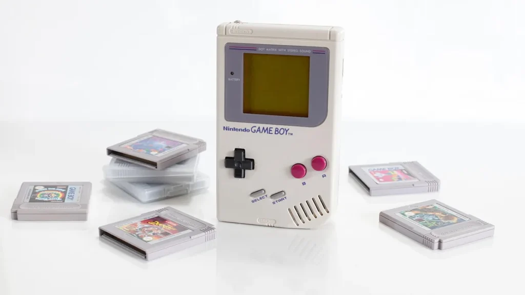 Grey Nintendo Game Boy console with red buttons stands on a white surface surrounded by game cartridges.
