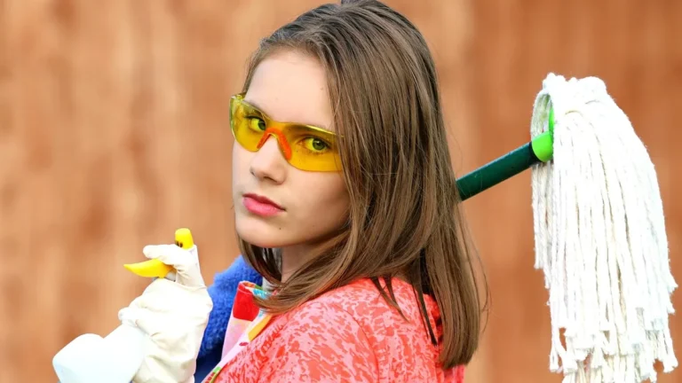 Young woman in yellow safety goggles holding a spray bottle with a mop over her shoulder.