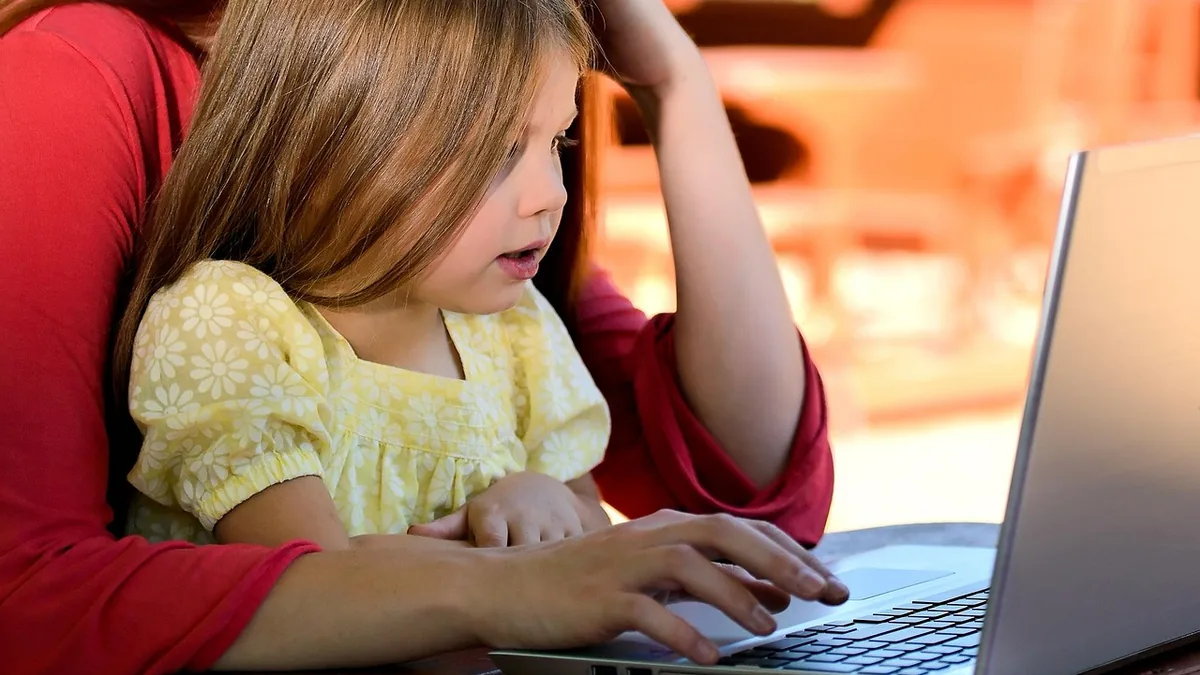 A young girl in a yellow dress looks surprised while sitting on an adult's lap, using a laptop.