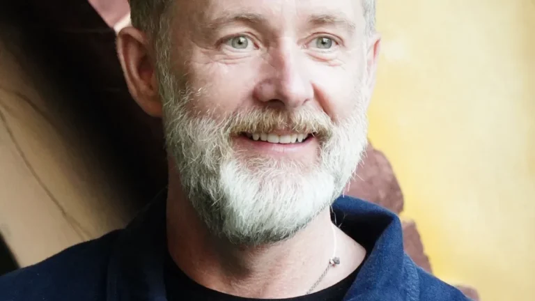 Close-up portrait of a smiling man with a grey beard, green eyes, and a blue shirt.