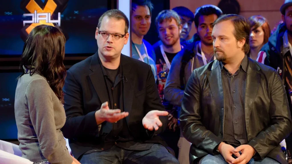 Greg Zeschuk speaks in a gaming interview alongside Ray Muzyka, with a PLAY logo in the background.