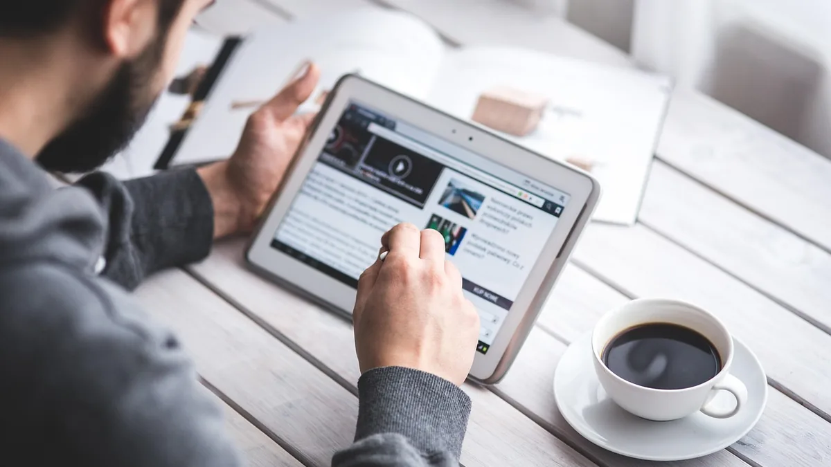 Man viewing news on a tablet while holding a cup of coffee at a wooden table.