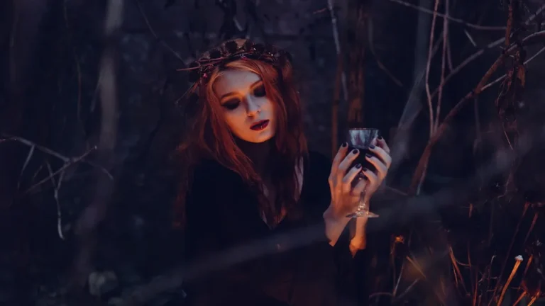 Red-haired woman wearing a crown holds a dark chalice in a shadowy, mysterious forest setting.