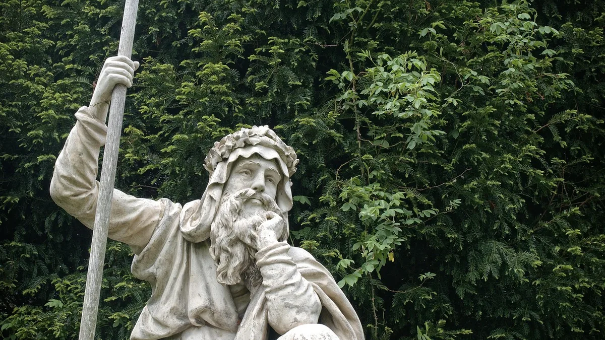A white stone statue of a bearded druid holding a staff against a green leafy background.