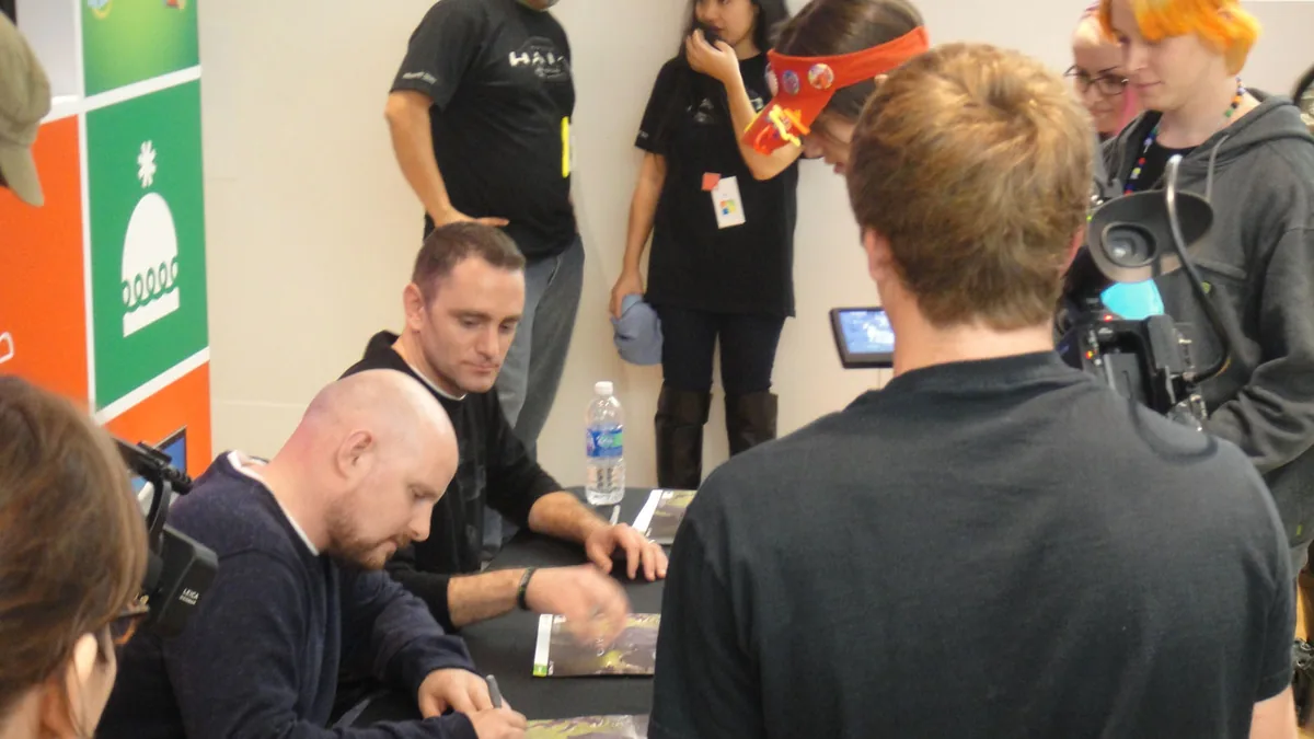 People gather around a table where developers are signing posters at a gaming event.