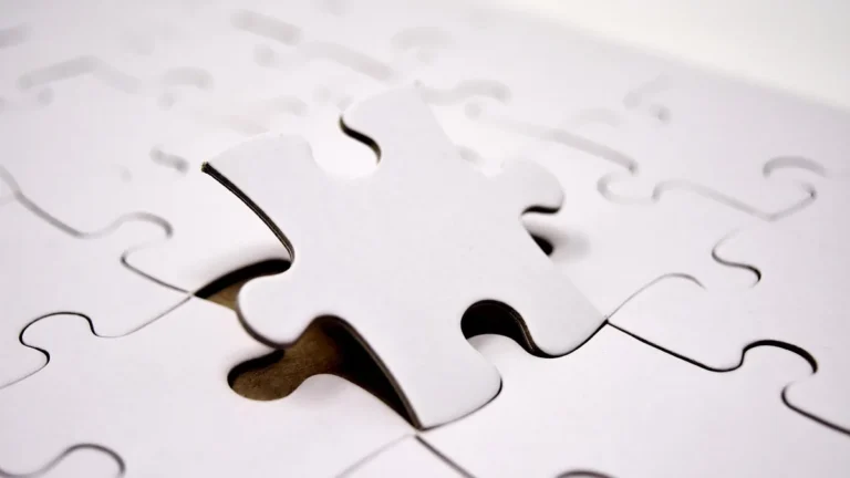 Close-up of a white puzzle piece fitting into a matching slot on a white puzzle board.