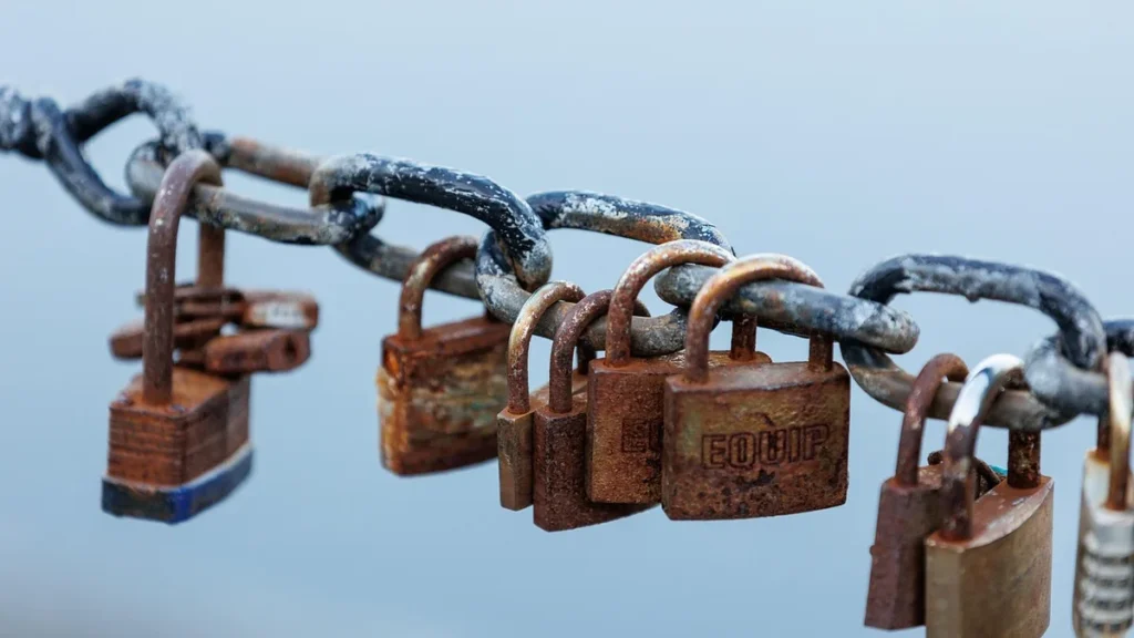 Rusty padlocks, including one engraved with the word EQUIP, locked onto a heavy metal chain.