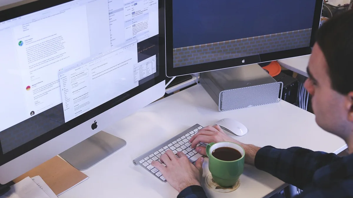 Man typing on a keyboard with dual monitors and holding a green coffee mug at a clean white desk.