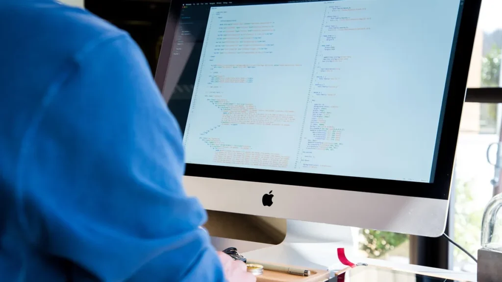 A person in a blue shirt viewed from behind writes code on a large desktop monitor showing lines of programming.