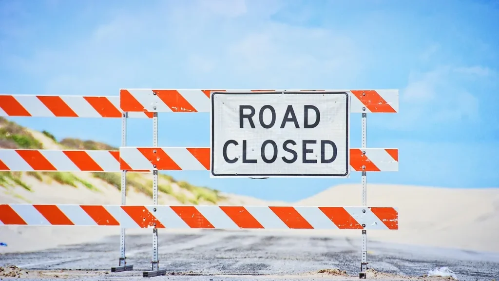 A white sign reading 'ROAD CLOSED' on an orange and white striped construction barrier blocking a sandy road under a blue