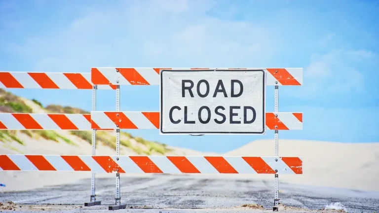 A white sign reading 'ROAD CLOSED' on an orange and white striped construction barrier blocking a sandy road under a blue
