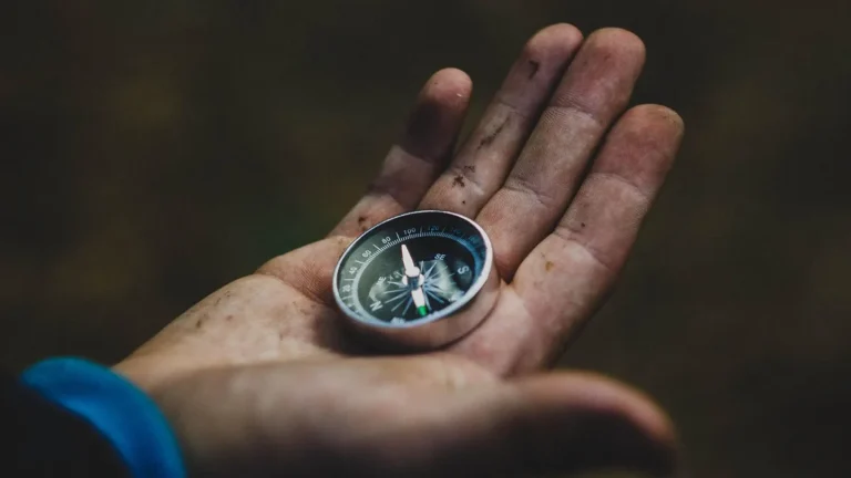 A dirty hand holding a small silver compass against a dark, blurry background.