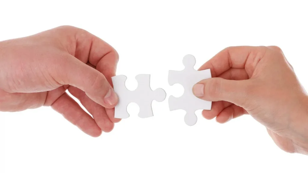 Two hands holding white jigsaw puzzle pieces about to connect, symbolizing teamwork and collaboration.