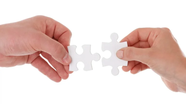 Two hands holding white jigsaw puzzle pieces about to connect, symbolizing teamwork and collaboration.