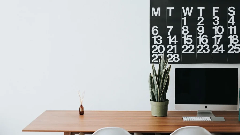 Minimalist wooden desk with a computer, potted snake plant, and a large black calendar on a white wall.