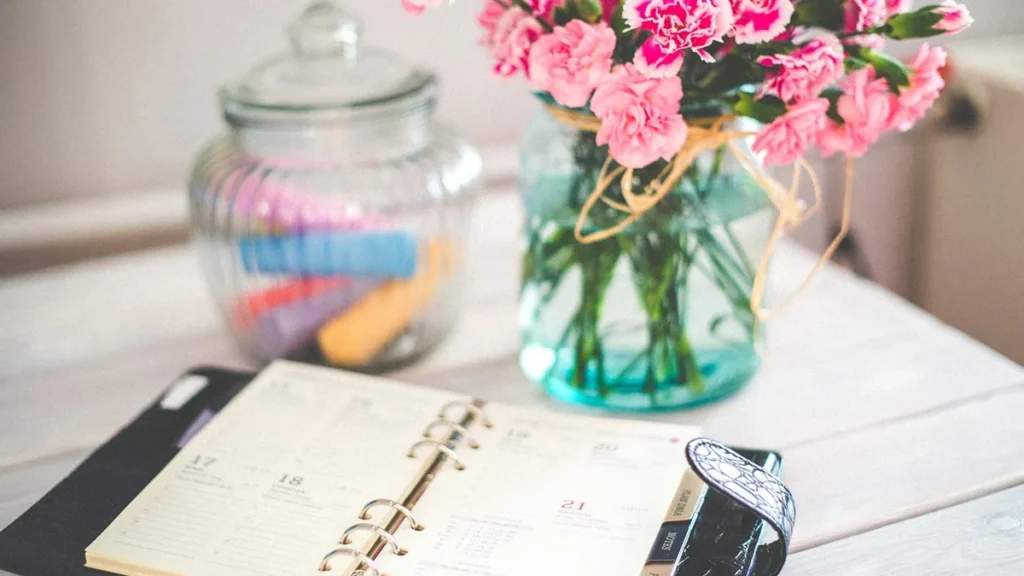 Open weekly planner on a white wooden table next to pink flowers in a blue glass jar.