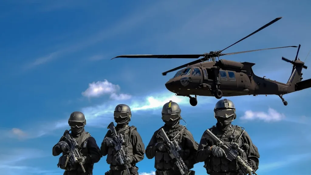 Four soldiers in black tactical gear holding rifles stand in front of a black helicopter in a blue sky.