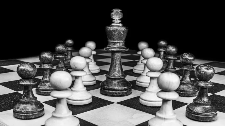 Black and white close-up of a large wooden king chess piece standing among pawns on a chessboard.