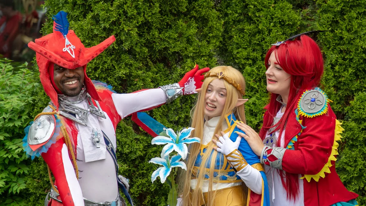 Three cosplayers dressed as colorful Zelda characters pose playfully against a lush green hedge background.