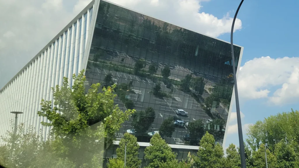 A modern corporate building with a massive glass facade reflecting a parking lot and green trees under a partly cloudy sky.