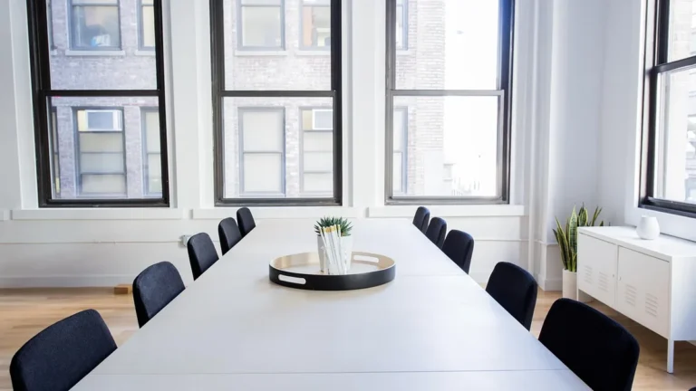Empty corporate boardroom with a long white table, black chairs, and large windows.