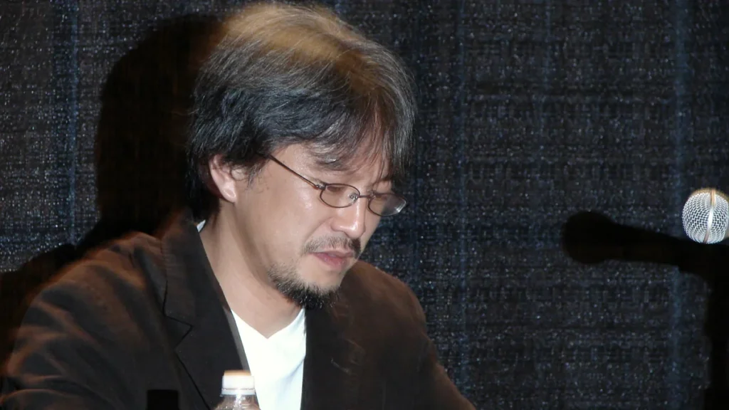 Portrait of Eiji Aonuma wearing glasses and a dark jacket, looking downward against a dark backdrop.