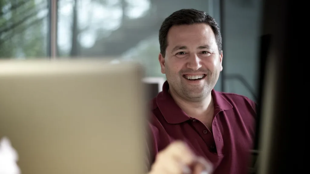 Smiling man in a maroon polo shirt sitting behind a laptop in an office setting.