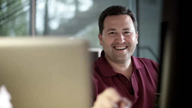 Smiling man in a maroon polo shirt sitting behind a laptop in an office setting.