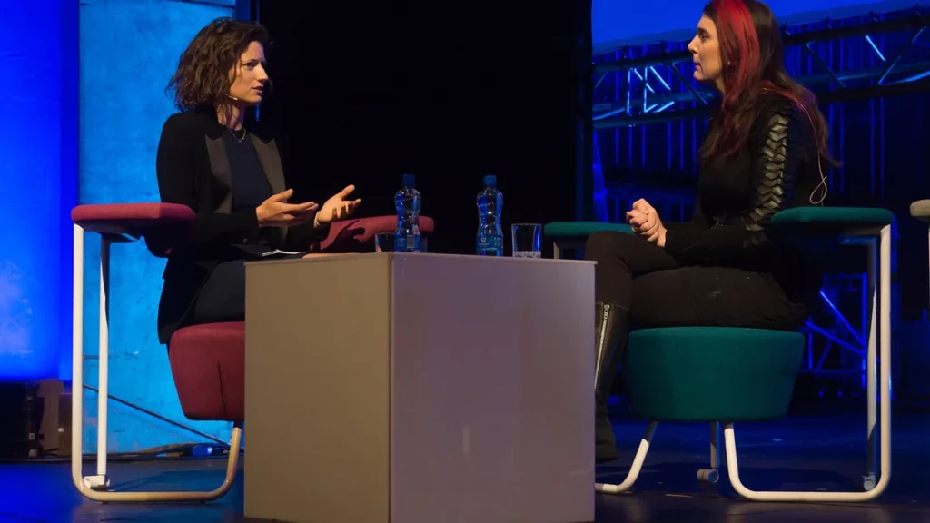 Two women speaking on a colorful stage panel, seated on red and teal chairs around a white table.