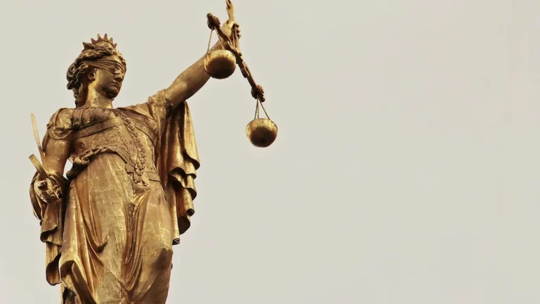 Golden statue of Lady Justice holding scales and sword against a plain light sky background.