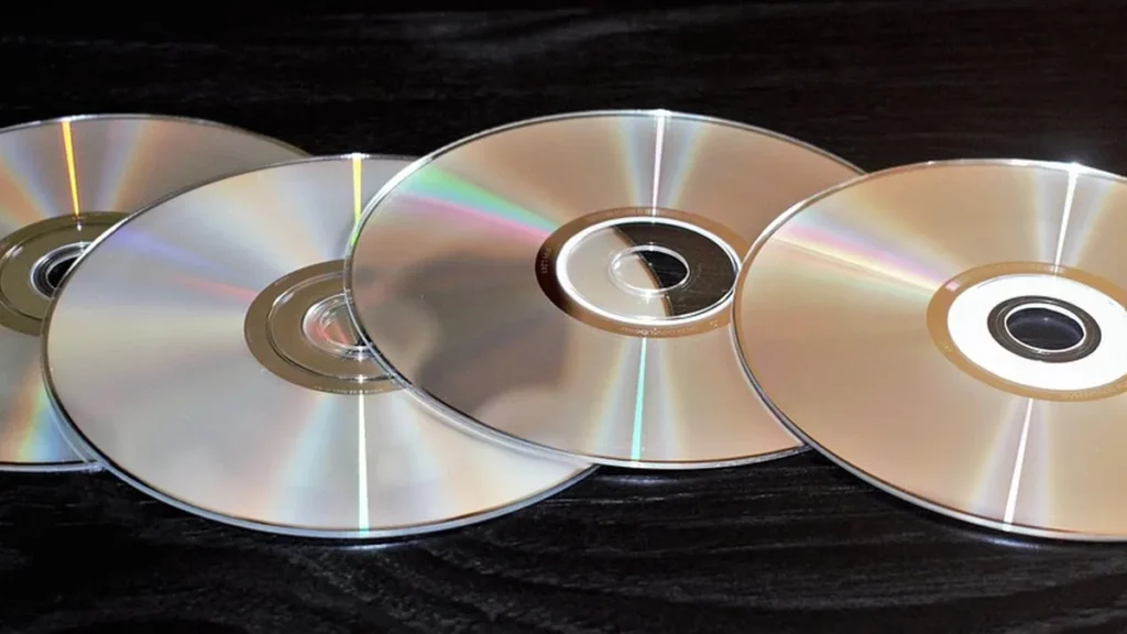 Four shiny optical discs overlapping on a dark wooden surface reflecting rainbow colors.