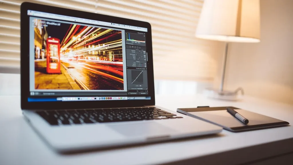 Laptop displaying photo editing software on a desk with a graphics tablet and stylus under a warm desk lamp.