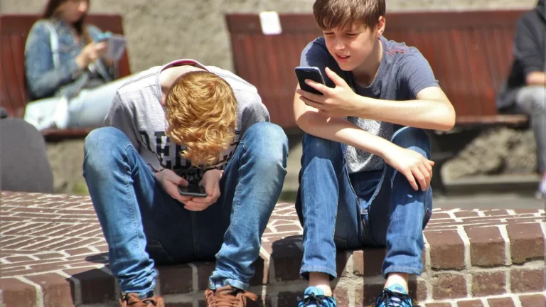 Two teenage boys sitting on a brick ledge, focused on their smartphones on a sunny day.
