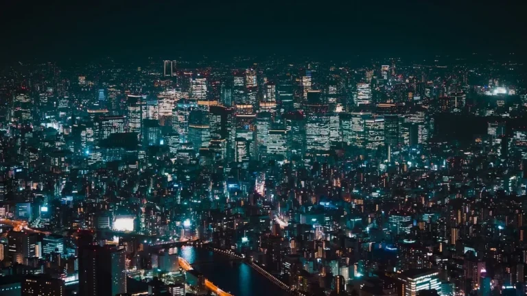 Night view of a sprawling city glowing with teal and orange lights, with a river and bridge below.