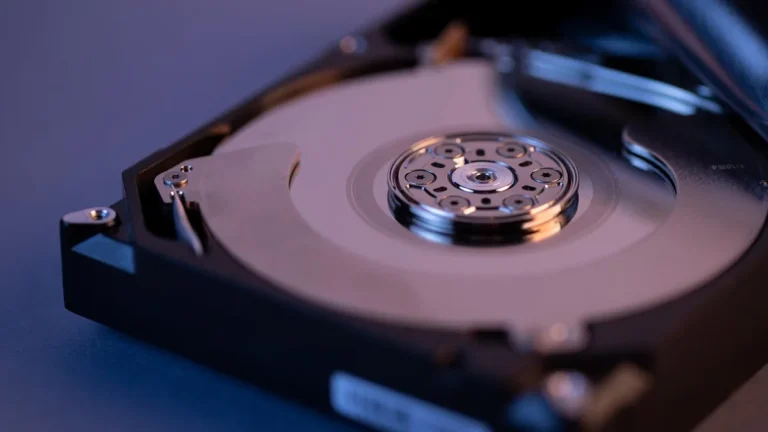 An opened hard disk drive showing the shiny metallic platter and spindle under blue and purple light.