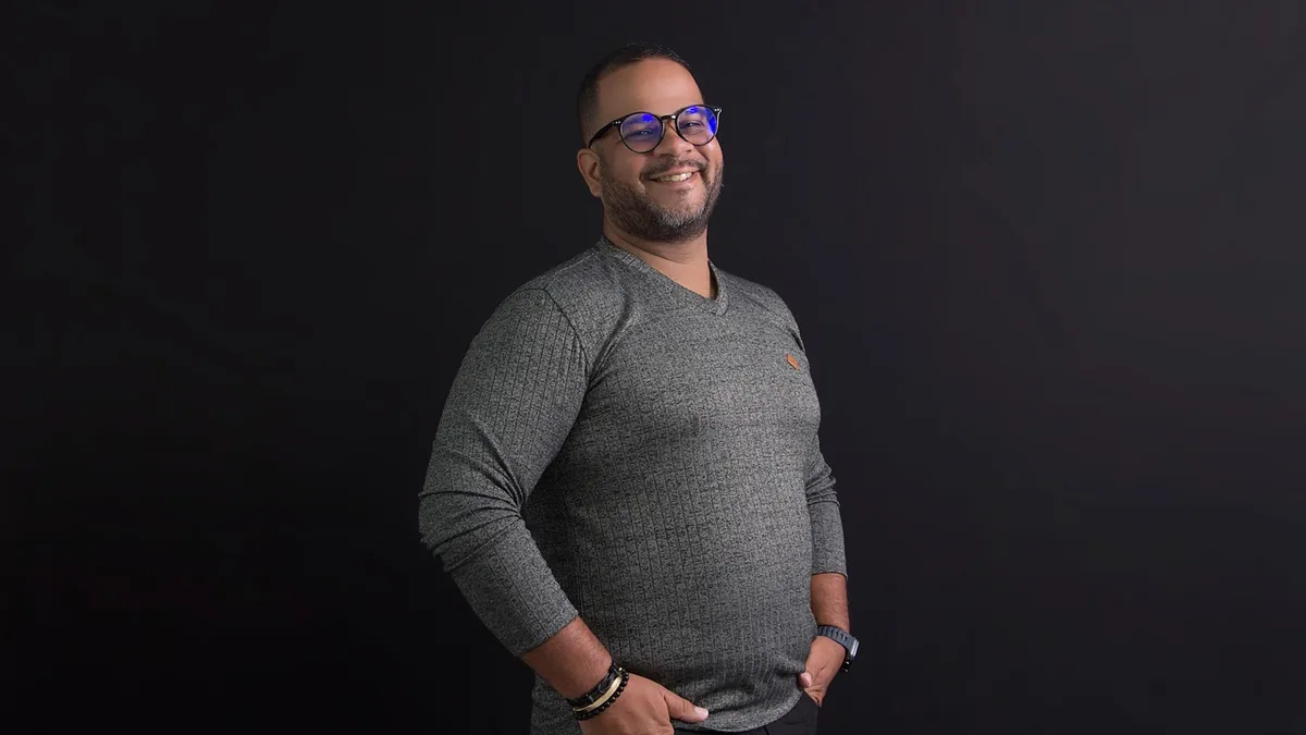 Smiling man with glasses in a grey shirt standing against a dark background.