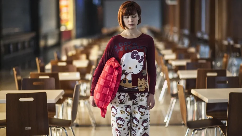 A woman in red Hello Kitty pajamas carrying a red pillow walks through empty canteen tables.