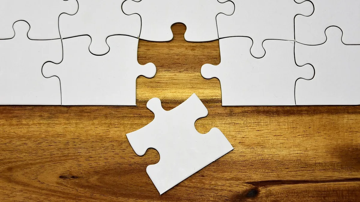 A single white puzzle piece sits on a wooden table below a row of white puzzle pieces with a missing gap.