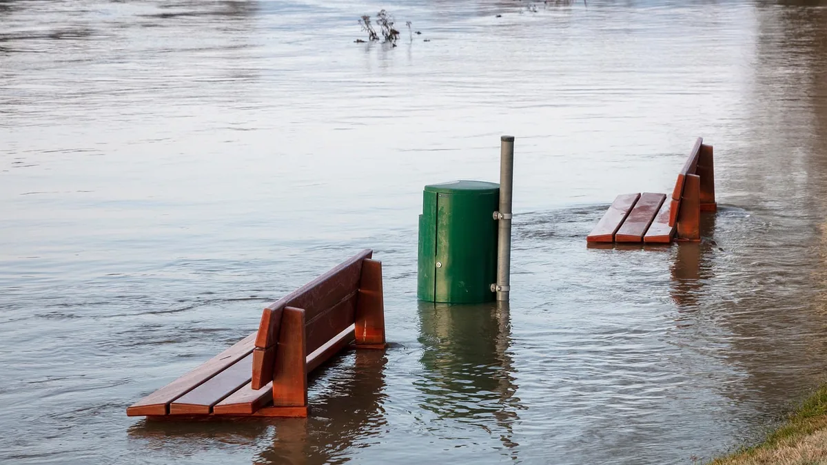 Wooden benches and a green trash can partially submerged in calm floodwaters.