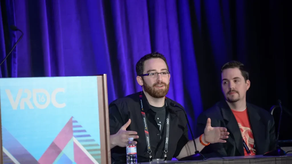 Two developers present on stage during a panel at the VRDC conference, behind a colorful VRDC 16 podium.
