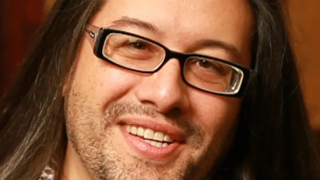 Close-up portrait of video game designer John Romero smiling, wearing black-rimmed glasses and long dark hair.