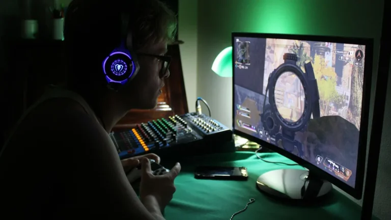 A gamer wearing a glowing purple headset plays a shooter game on a monitor in a green-lit room.