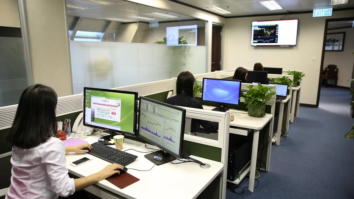 Office workers monitor financial charts on multiple computer screens in a corporate office setting.