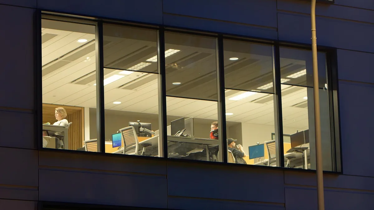 Office workers at desks with computer monitors, seen through a large window at night.