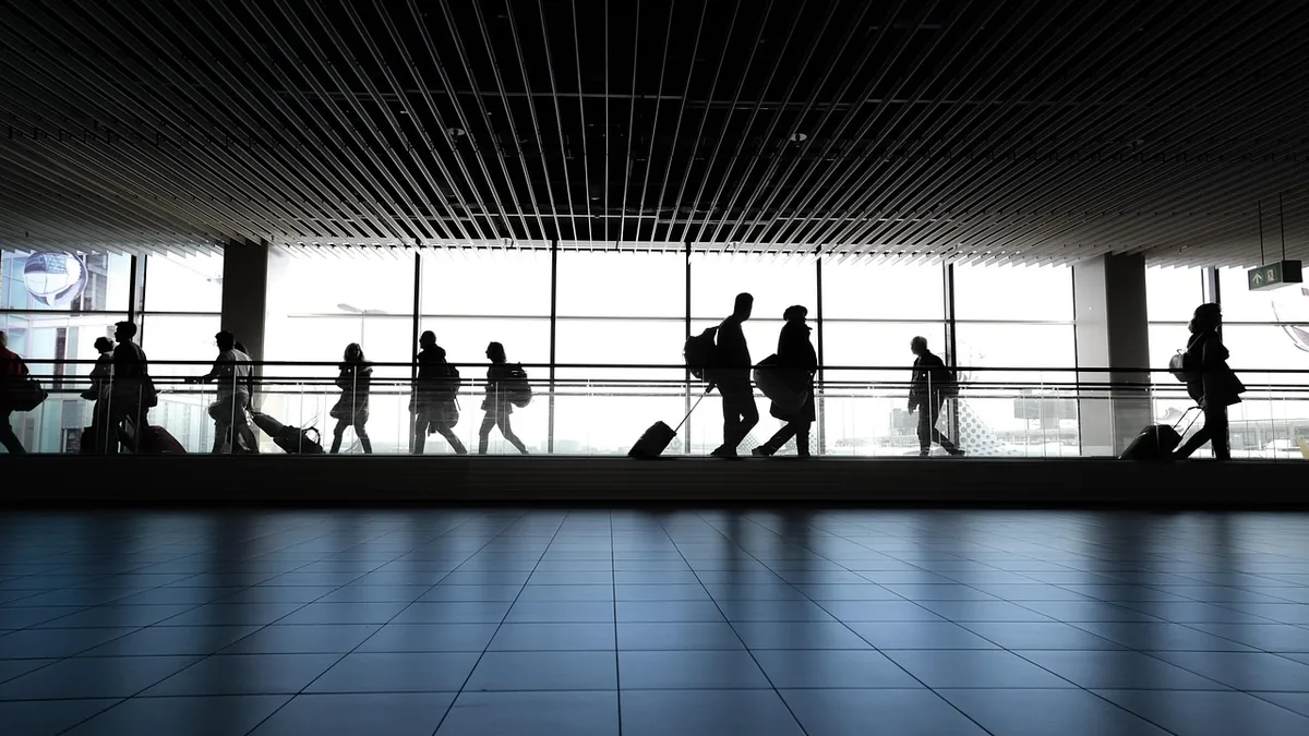 Silhouettes of travelers walking along a bright airport corridor under a slatted ceiling.